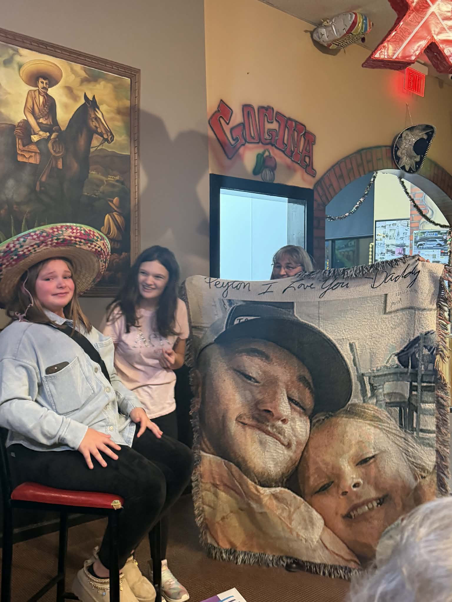 Two women sitting in a room with a large cutout of a man and child, surrounded by decor including a painting of a man on a horse and a Cacti bar sign.